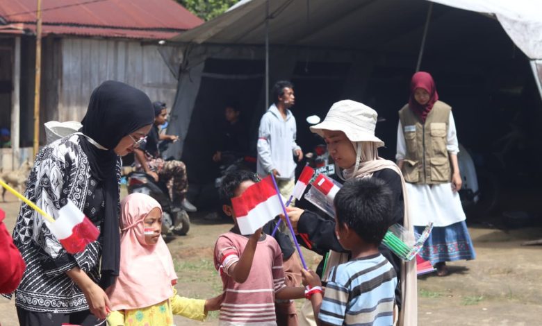 Relawan membagikan bendera kecil kepada anak-anak di pelosok Desa Tompu, Sigi, dengan latar tenda sederhana dan warga setempat.